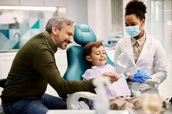 Father holding a mirror for his happy, young son sitting in a dental chair while dentist happily watches