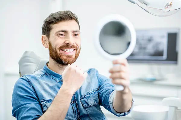 Bearded patient checking out his handsome smile in a mirror while sitting in a dental chair