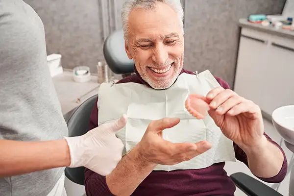 Elderly male patient smiling and examining a set of dentures during a consultation at a dental clinic.