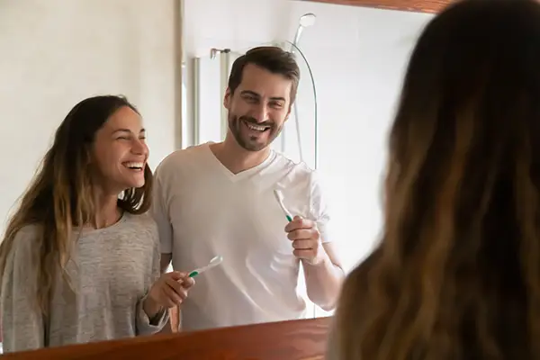 A smiling young couple in the bathroom mirror practicing good dental hygiene together.